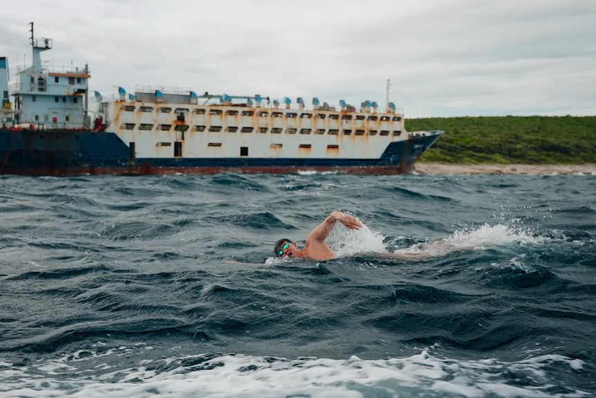 A male swimmer in choppy seas with a large ship nearby