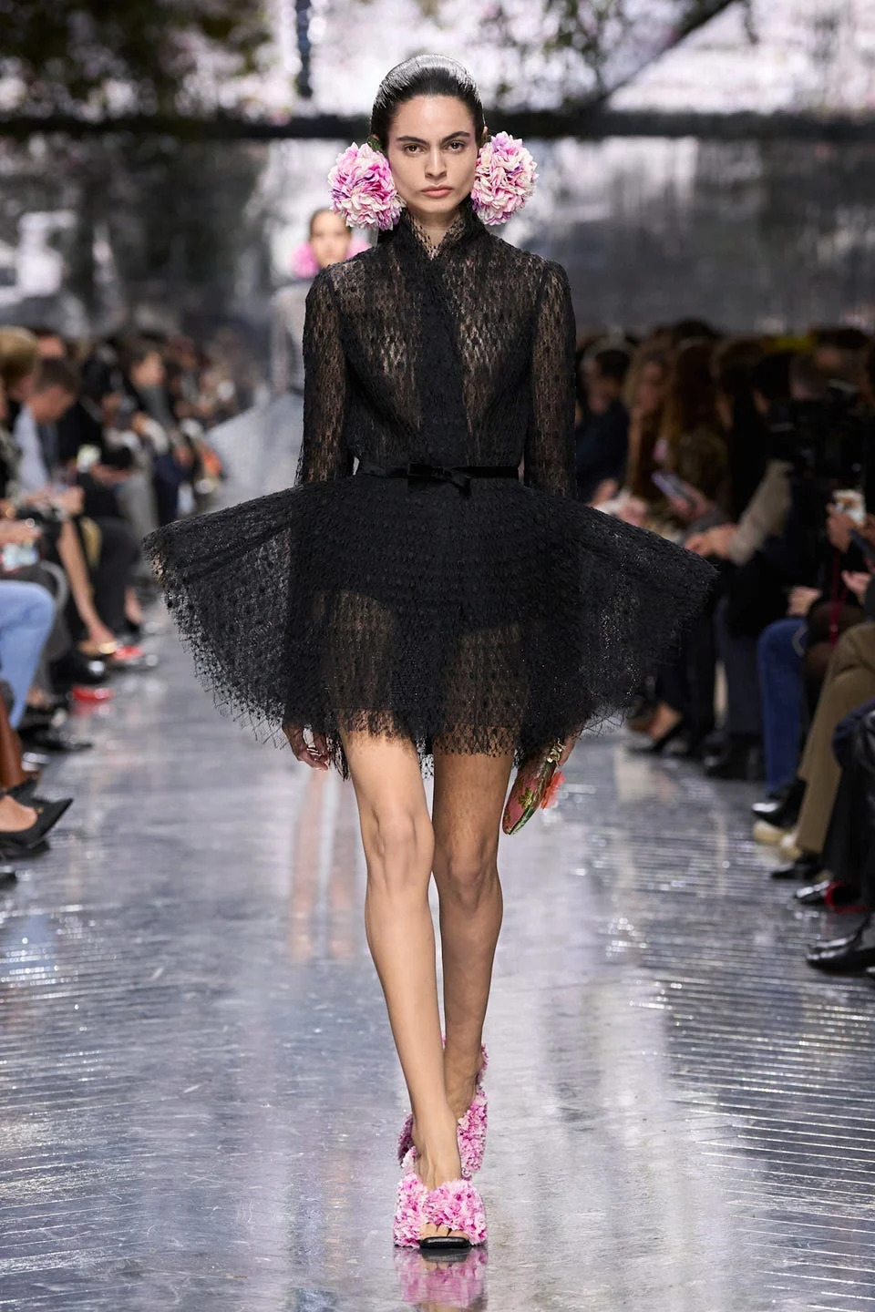 Model walking down runway in a black dress with floral accents.