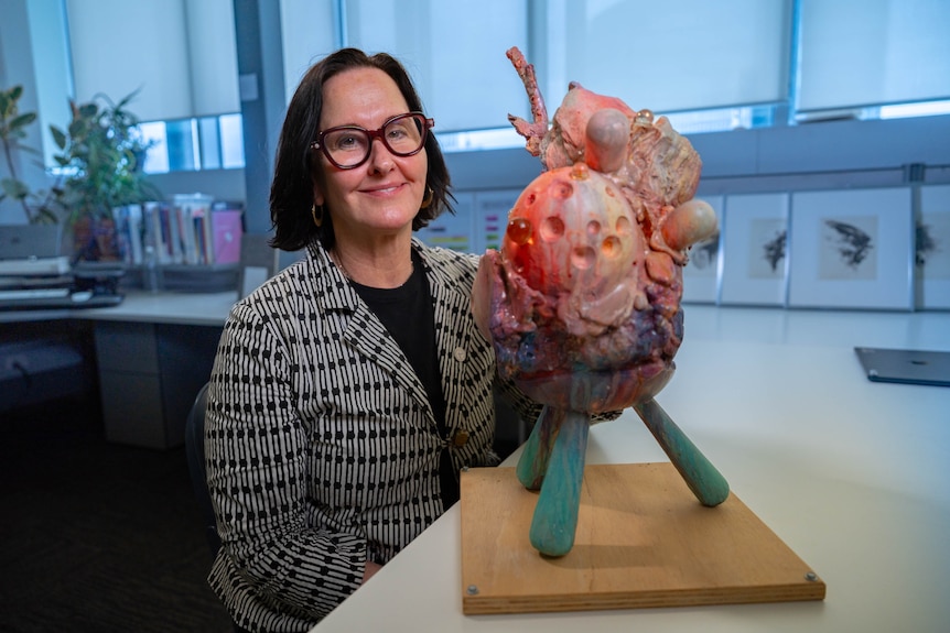 A woman with black hair and red glasses wearing a black and white suit sits beside a colourful sculpture of the heart.