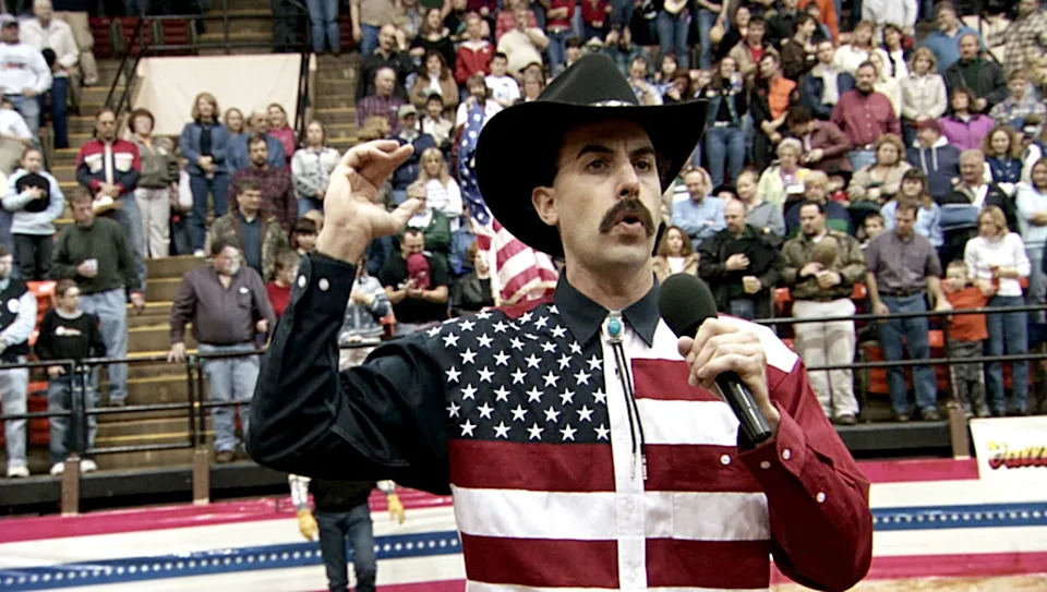 Man in cowboy hat with mustache holds microphone, wearing American flag shirt, speaking in crowded arena, U.S. flag visible behind