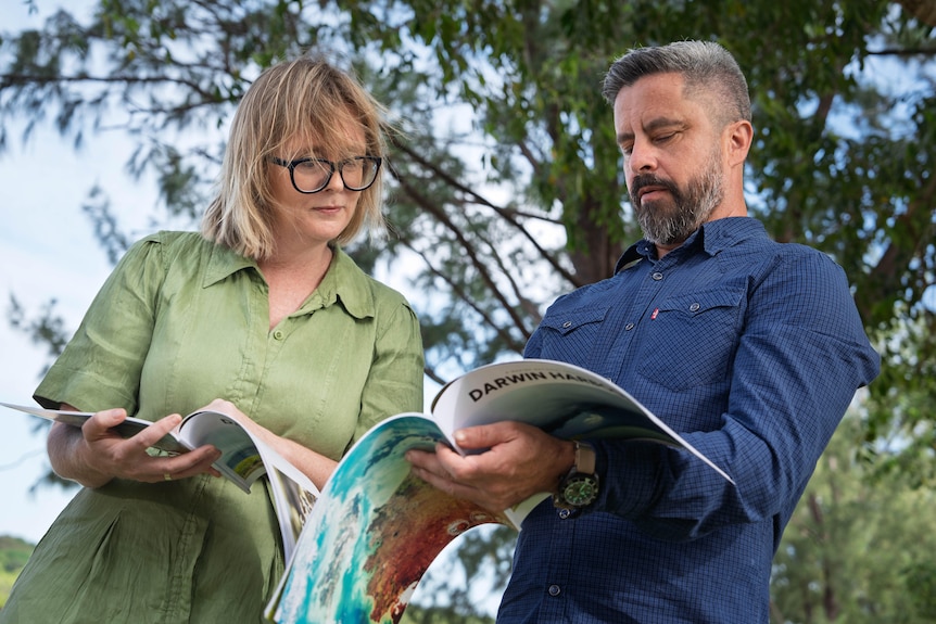 Two people standing side-by-side on a beach, looking over a report together.