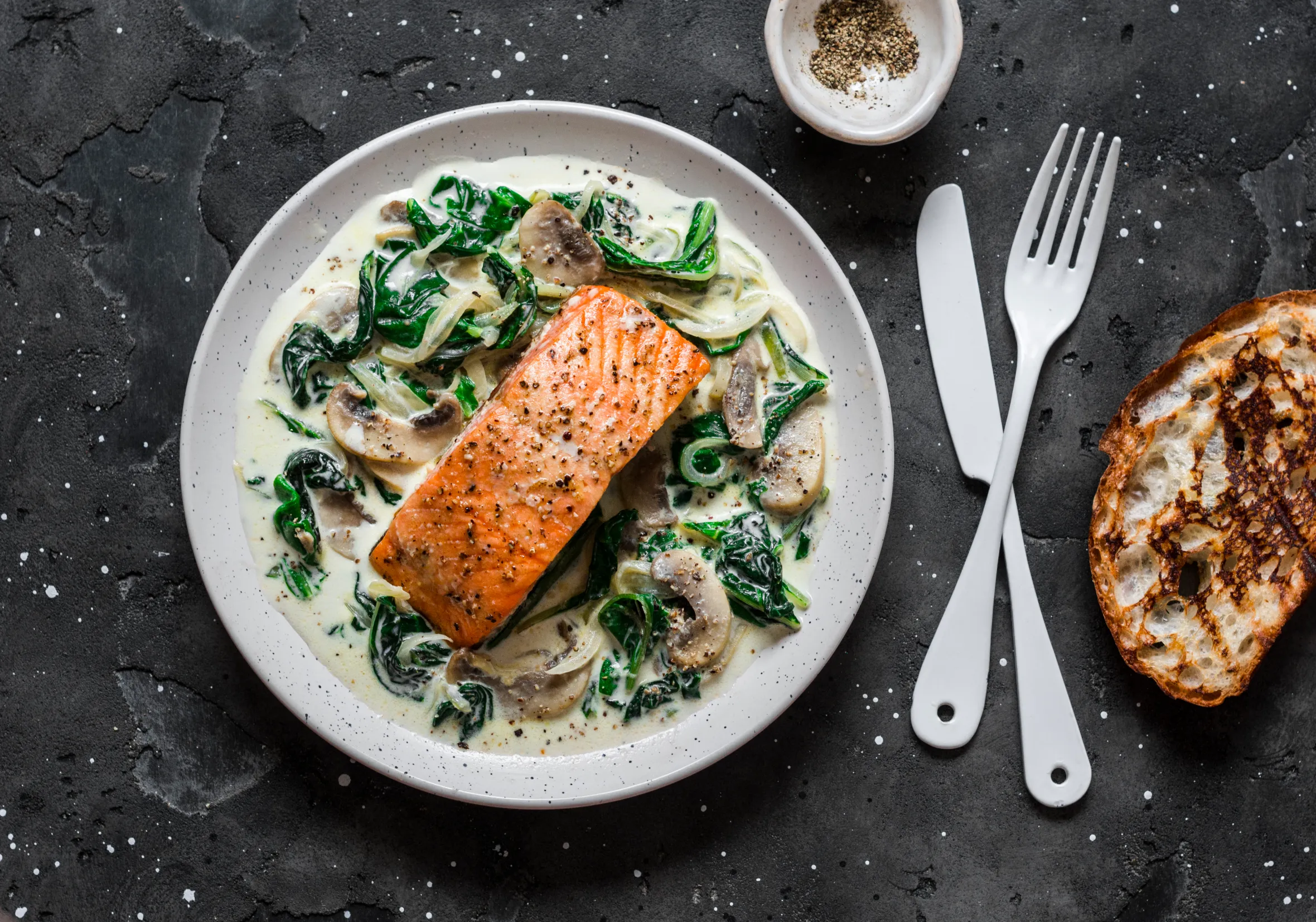 Baked salmon with creamy spinach mushroom sauce on a plate, served with a slice of toasted bread, fork, and knife.
