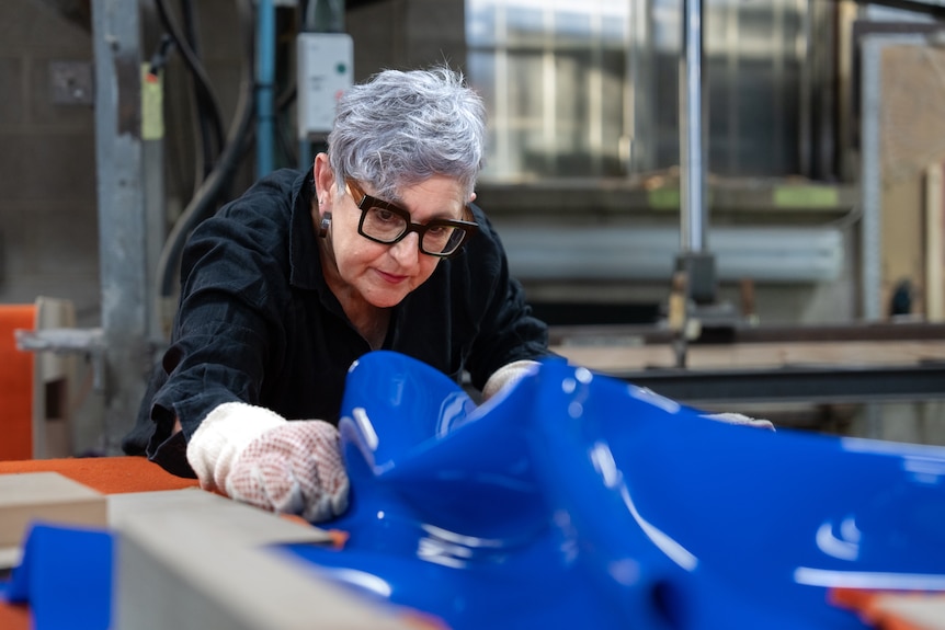 A woman with greying hair, wearing thick white gloves, holds a crumbled blue sheet of plastic.