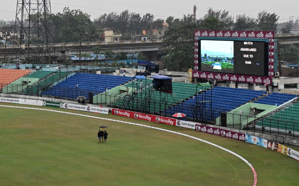 Men carrying an umbrella walk past a digital screen after the second T20 cricket match between Bangladesh and New Zealand was washed out owing to rain at the Bir Sreshtho Flight Lieutenant Matiur Rahman Stadium in Chittagong on April 29, 2026.