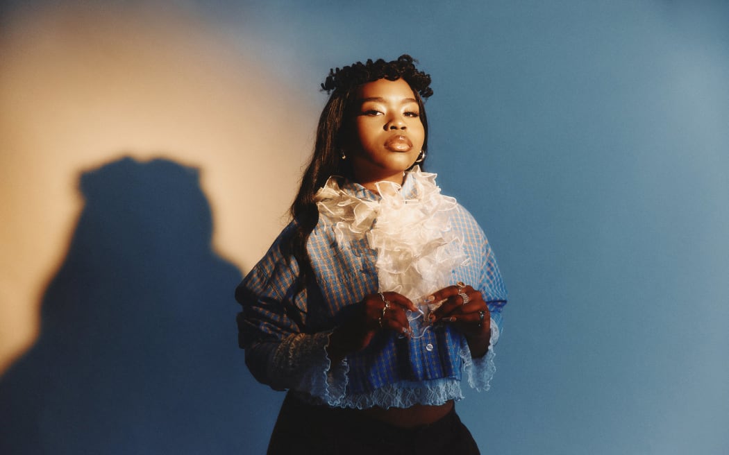 This is a photo of Tusekah. It looks like a studio shot. She is stood in front of a white background that appears blue and orange because of the lighting. Her shadow is visible on the wall behind her. She is looking towards the camera in a kind of self-assured way. She is wearing a blue stripey shirt with a big white frilly ruffle.