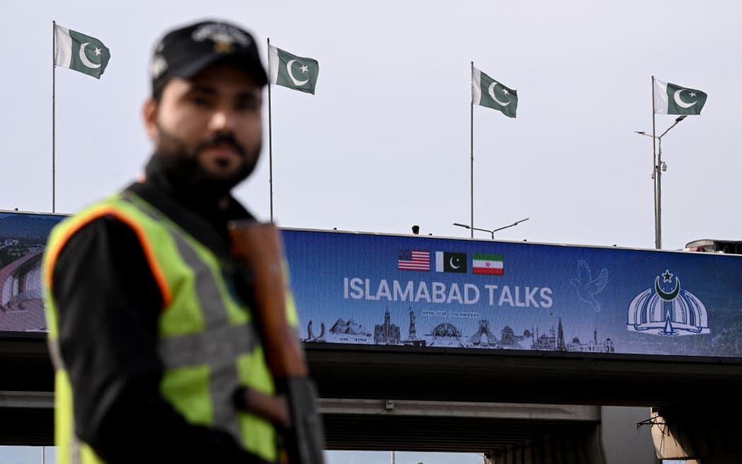 A policeman stands guard in front of a digital screen displaying news of US–Iran peace talks along a road in Islamabad on April 10, 2026. A cloud of uncertainty hung April 10 over the scheduled start of talks in Pakistan between the United States and Iran, with no announcement yet on the arrival of negotiators and both sides accusing the other of failing to properly implement a fragile ceasefire. (Photo by Farooq NAEEM / AFP)