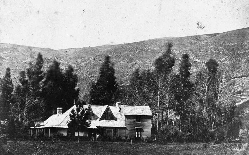 A view of the house owned by Thomas Mason in Taita, Lower Hutt, circa 1890s. The house is situated at the foot of a hill and is surrounded by tall trees. An unidentified man and woman (possibly the Thomas and Jane Mason) stand in front of the house.