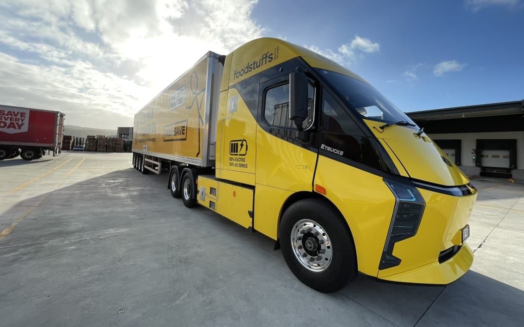 A bright yellow large electric truck and trailer unit with Foodstuffs branding is shown driving slowly out of a freight depot