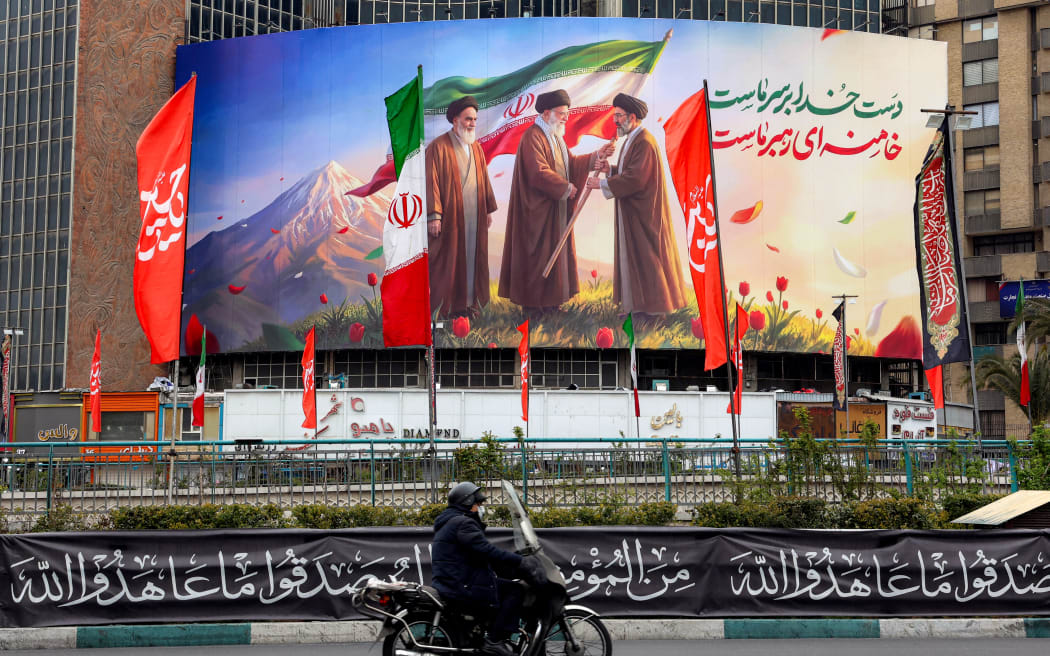 A man rides a motorcycle past a banner displayed at Valiasr Square in central Tehran on March 10, 2026, depicting Iran's late supreme leader Ayatollah Ruhollah Khomeini (L) watching as his successor the late Ayatollah Ali Khamenei (C) hands over a national flag to his son and new supreme leader Mojtaba Khamenei.