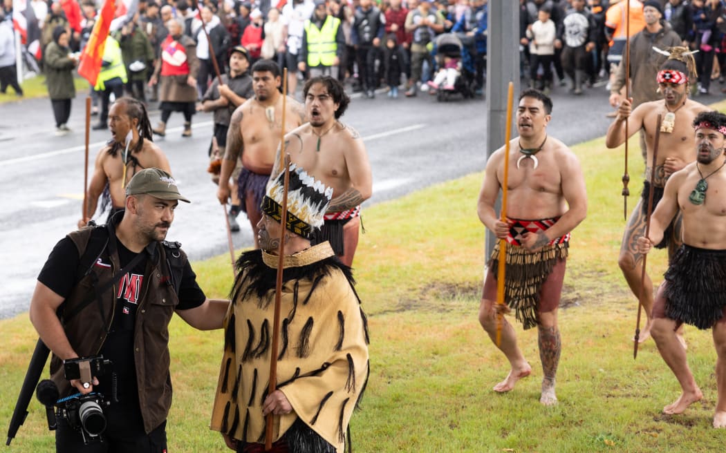 Whatanui is holding his camera and walking at the front of the Hikoi. He is in conversation with one of the leaders who is wearing a korowai.
