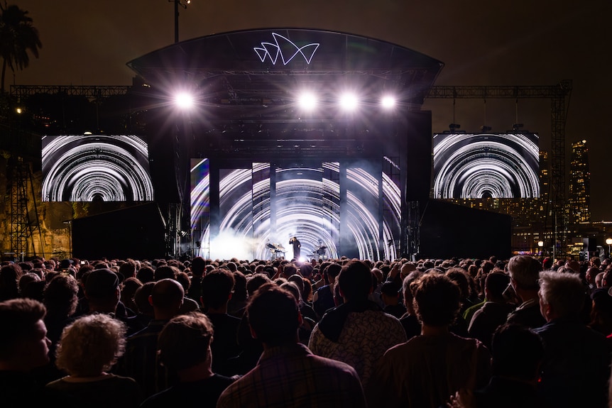 Wide shot of Sydney Opera House forecourt stage behind audience, screens show large b&w rings and small Thom Yorke centrestage