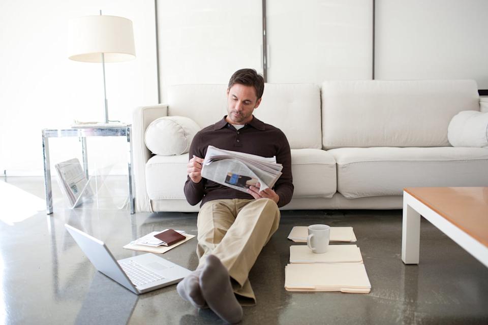 A man sitting against the couch, reading the newspaper.