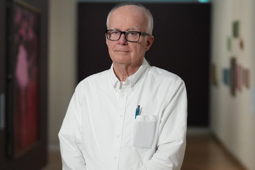 A man with white hair and glasses stands in an art gallery looking serious.