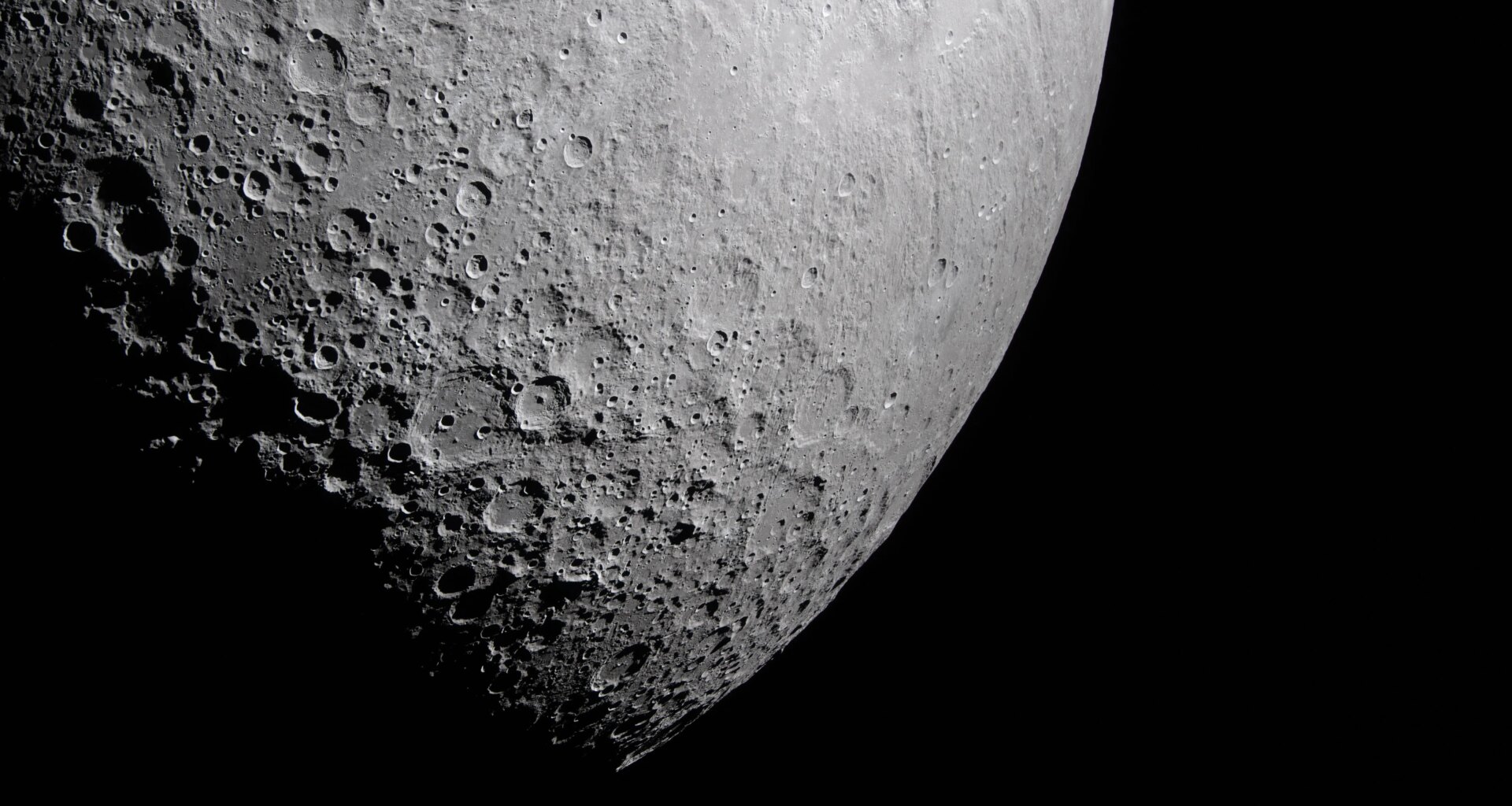 Part of the Moon stands out against the blackness of space. Its surface is marked with many craters of different sizes. The craters are mostly on the left and bottom. The upper right portion of the Moon's surface is much smoother.