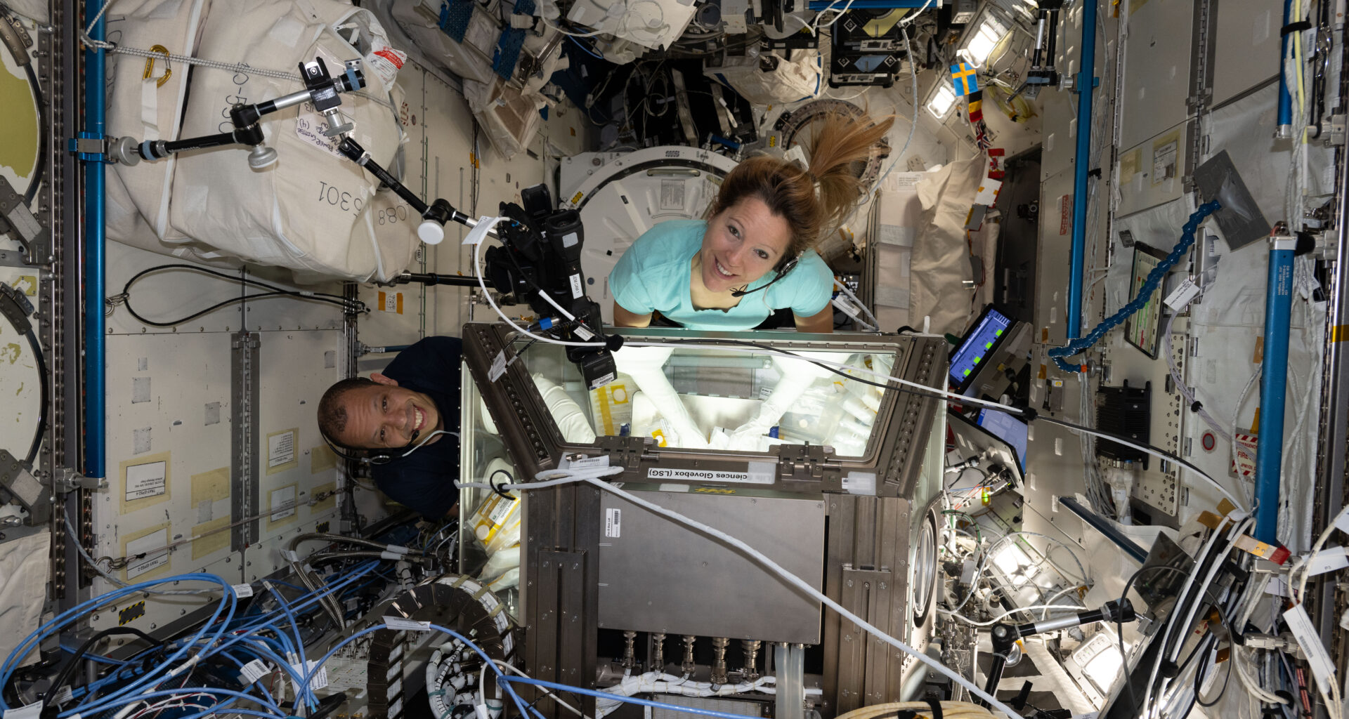 Two people look up at a camera. They each have their arms in sleeves that go inside a lit-up rectangular box. We can see their arms through the windows on each side of the box. There are wires all around them inside the International Space Station.