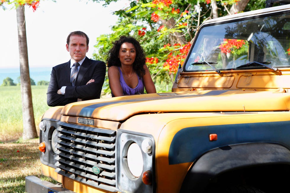 Ben Miller, Sara Martins, Death in Paradise ben miller and sara martins standing next to a vintage yellow land rover defender in death in paradise