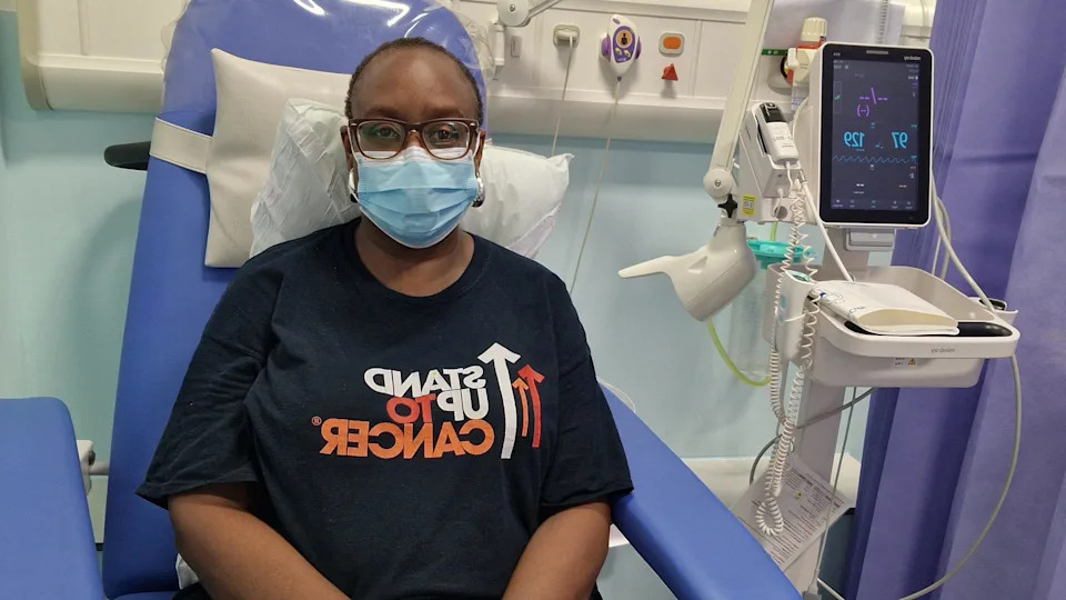 Simone Boothe wearing a "Stand up to cancer" t-shirt in a hospital, wearing a blue face mask.
