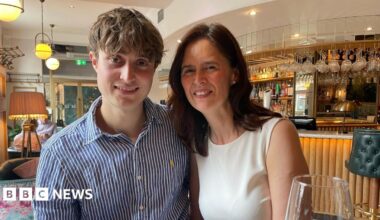 A woman in a red dress looks at the camera in front of a green bush and pink flowers.