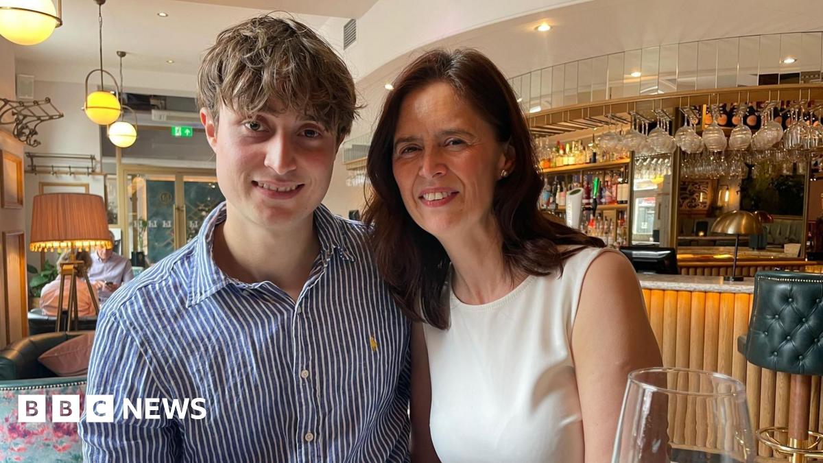A woman in a red dress looks at the camera in front of a green bush and pink flowers.