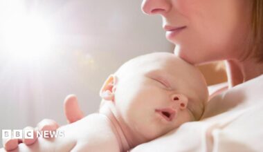A baby sleeping on his or her mother's chest. The mum's chin, nose and lips can be seen with her chin gently resting on the baby's head. She wears a light top and has a blonde bob.