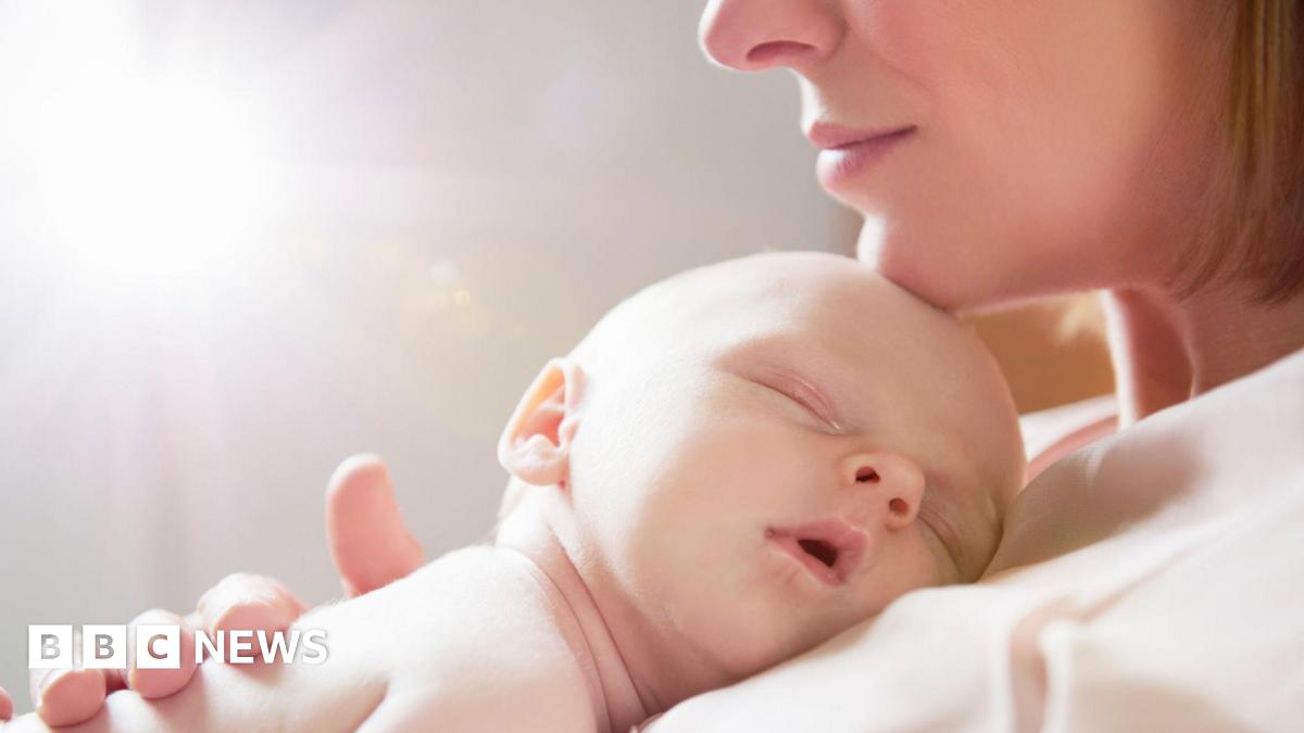A baby sleeping on his or her mother's chest. The mum's chin, nose and lips can be seen with her chin gently resting on the baby's head. She wears a light top and has a blonde bob.