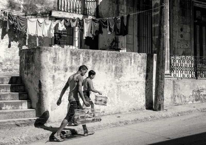 Two men walk down a street carrying boxes in front of a weathered building with a line of laundry hanging above them. The scene is in black and white, with bright sunlight casting strong shadows.