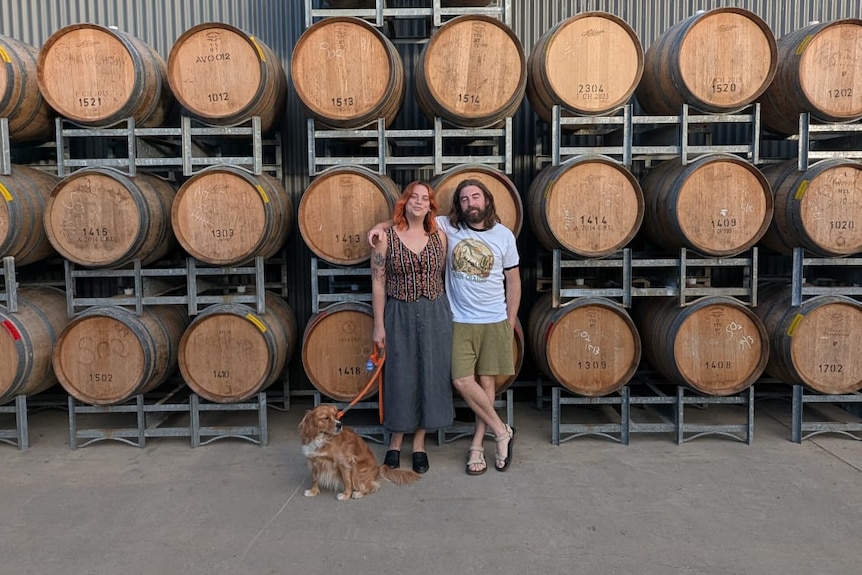 young man and woman stand in front of rows of wine barrels. they are holding a dog on a lead.