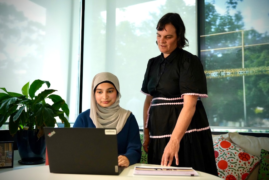 Hafsa is a teenager who wears a headscarf and types on a computer. Katherine wears a dress and watches over her shoulder.
