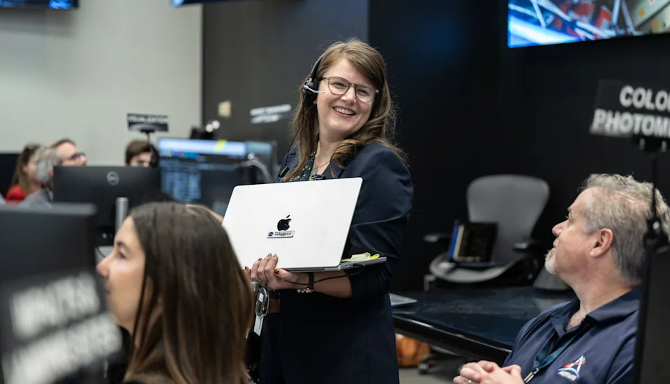 A woman in a control room, smiling and holding an open laptop, surrounded by colleagues working at computer stations