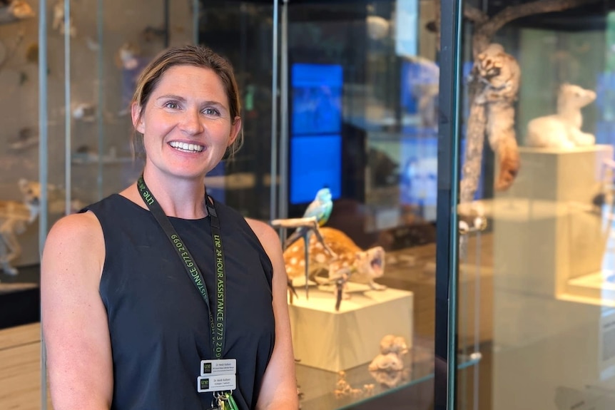 A smiling woman stands beside a glass cabinet containing taxidermied animals in a museum.  