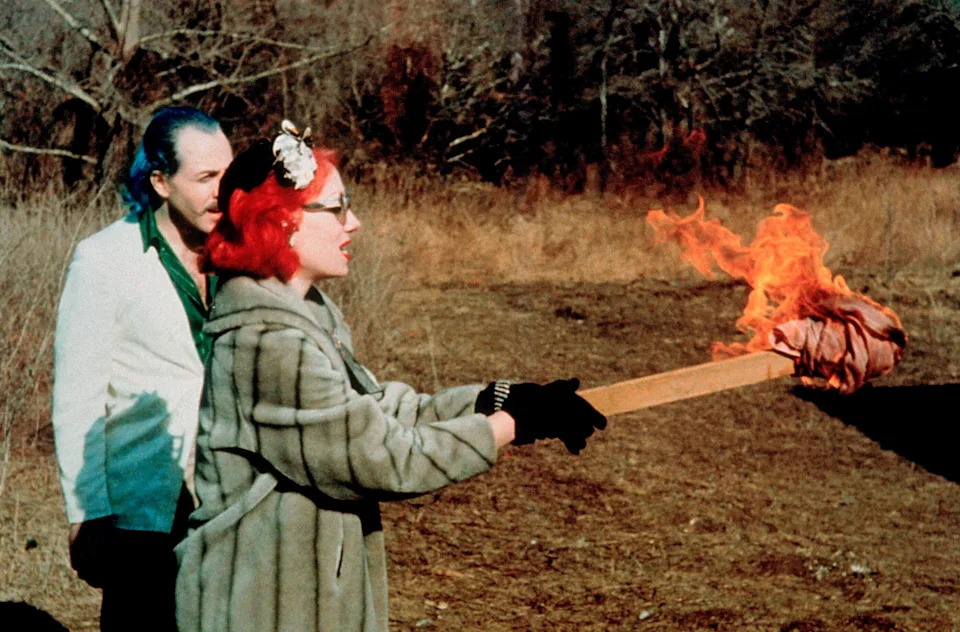 Two people outdoors. One is wearing a faux fur coat with a hair accessory, holding a flaming torch. The second person stands behind observing