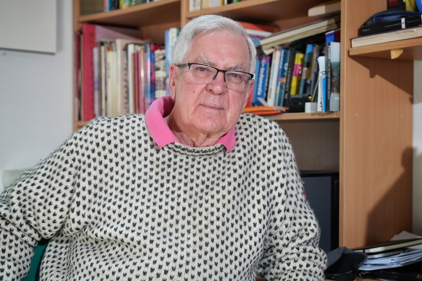 A man with white hair and spectacles sitting at a desk, with a bookshelf behind him.