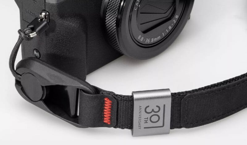 Close-up of a black camera with a lens attached, featuring a black strap with red stitching and a metal tag that reads "30th Anniversary.
