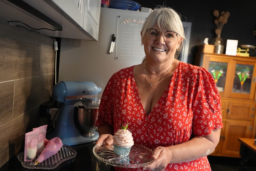 A fair-skinned woman in a red floral dress holds a cupcake on a glass plate in a kitchen.