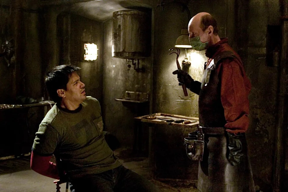 A person tied to a chair looks at another wearing a surgical mask and apron, holding a tool in a dimly lit, ominous room