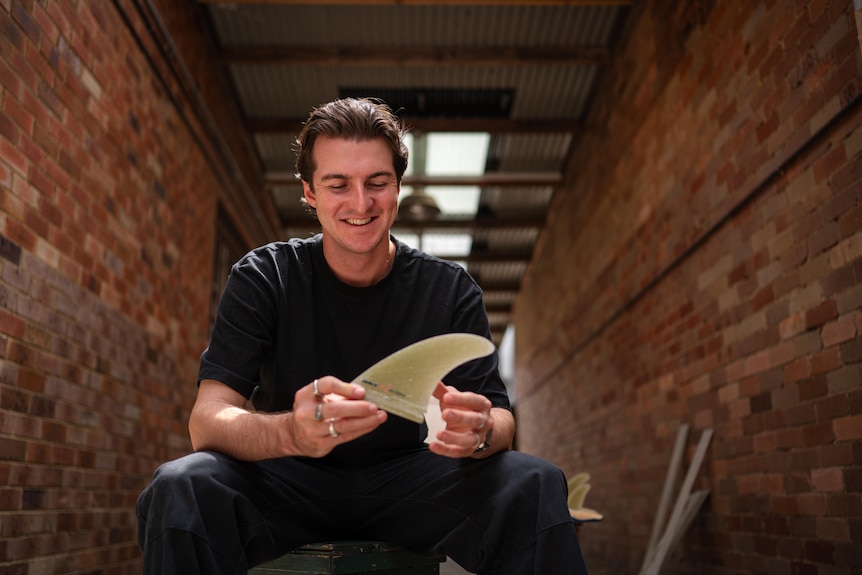 Man in black t-shirt smiling and holding a surfboard fin. 