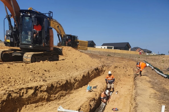 A digger next to a trench with three men in high vis out fits working. 