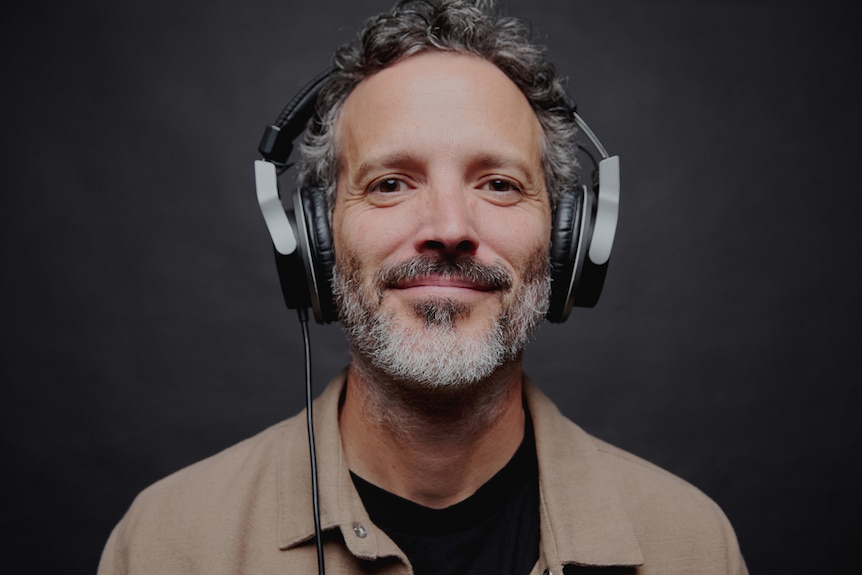Bret with grey-speckled beard smiles wearing silver pair of over-ear headphones, background is dark, wears beige shirt.