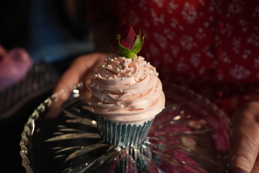 Pink frosted cup cake close up with white sprinkles and a rose on top. 