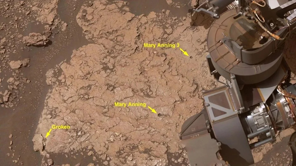 A close up of a reddish brown surface with a metal arm in the right top corner of the image. The image includes yellow arrows and labels of different sites of interest. .