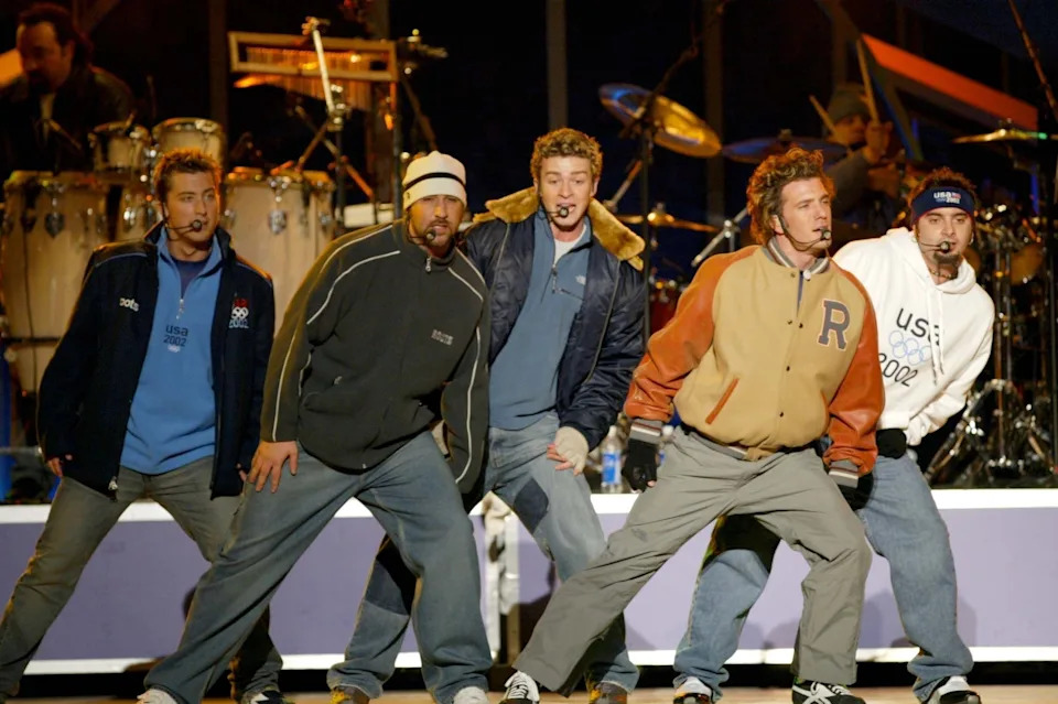 A group of male performers sing and dance on stage, wearing casual and sporty outfits, with a drum set in the background
