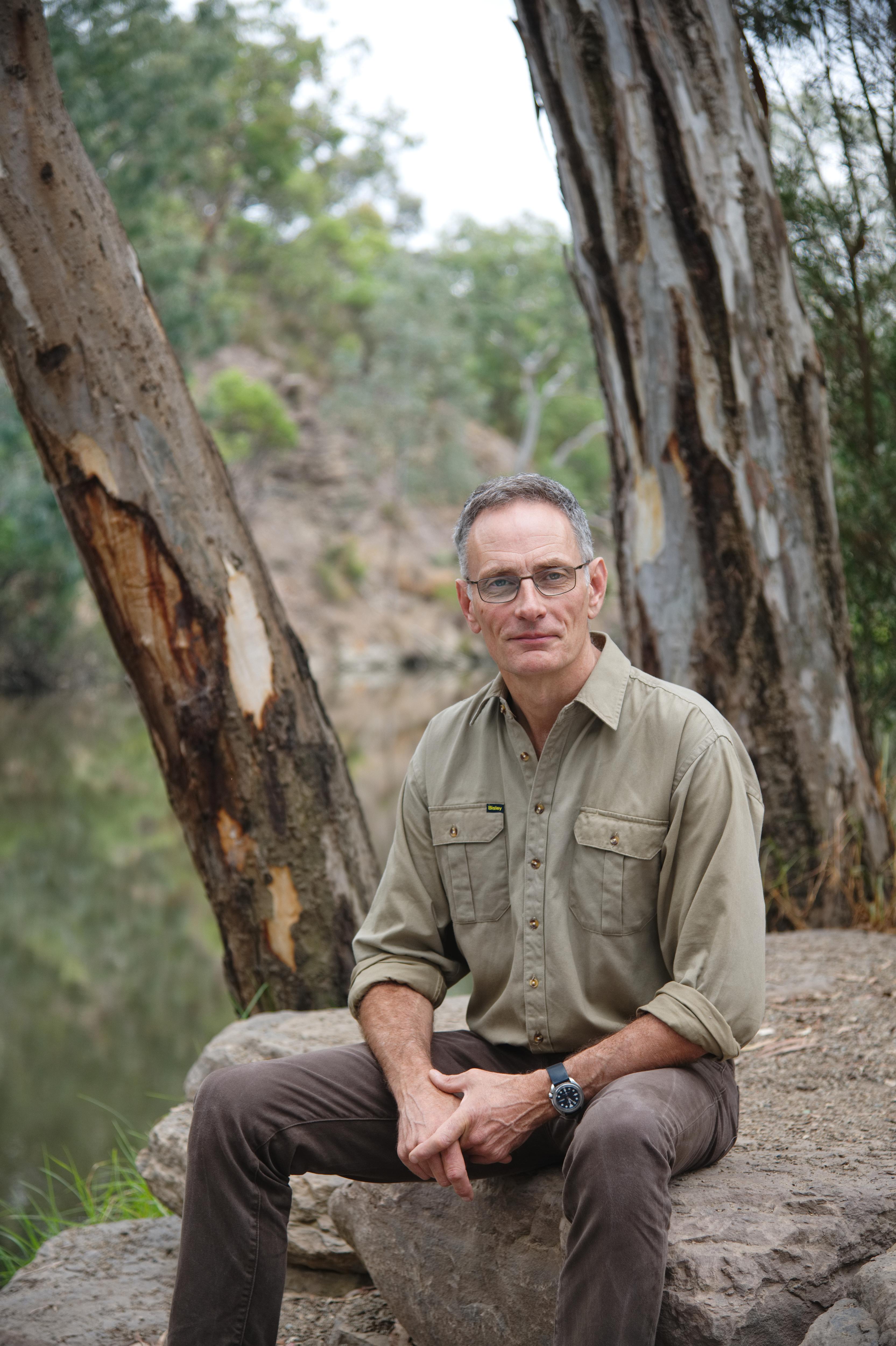 A man with grey hair and glasses in a khaki collared shirt and brown pants sits on a rock with his hands clasped in bushland.