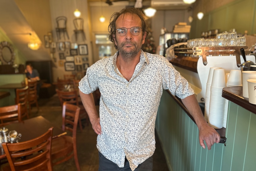 A man in a white shirt looks at the camera from within his cafe.