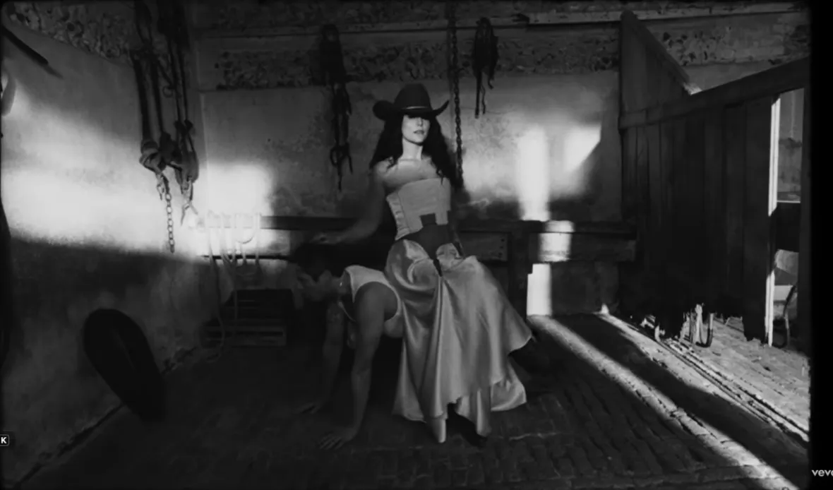 A woman in a cowboy hat and long skirt sitting on the back of another woman crawling on all fours.