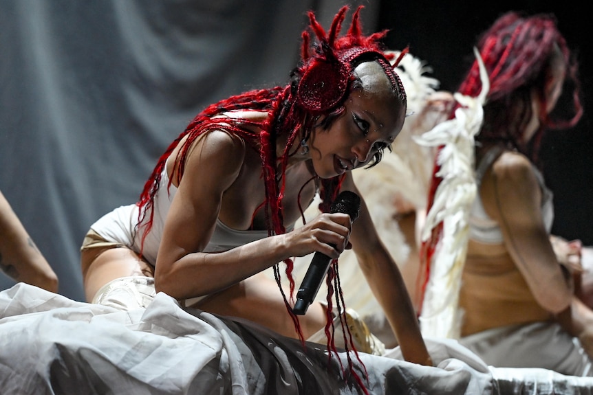 FKA twigs wearing white costume leans forward holding a microphone on a dimly lit stage, with dancers behind
