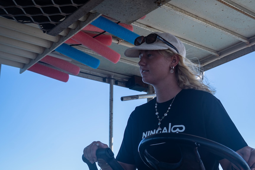 A blond woman wearing a hat stands at the controls of a boat.