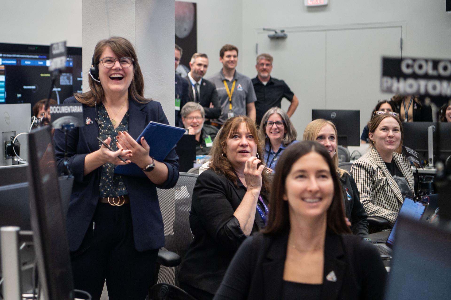 A large group of people sitting behind computer monitors smile at something just off camera to the right.