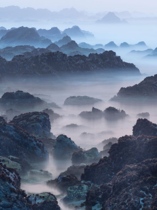 Misty landscape with dark, jagged rocks covered in moss, receding into the distance. Soft fog drifts among the rocks, creating a dreamlike, ethereal atmosphere with shades of blue and green.
