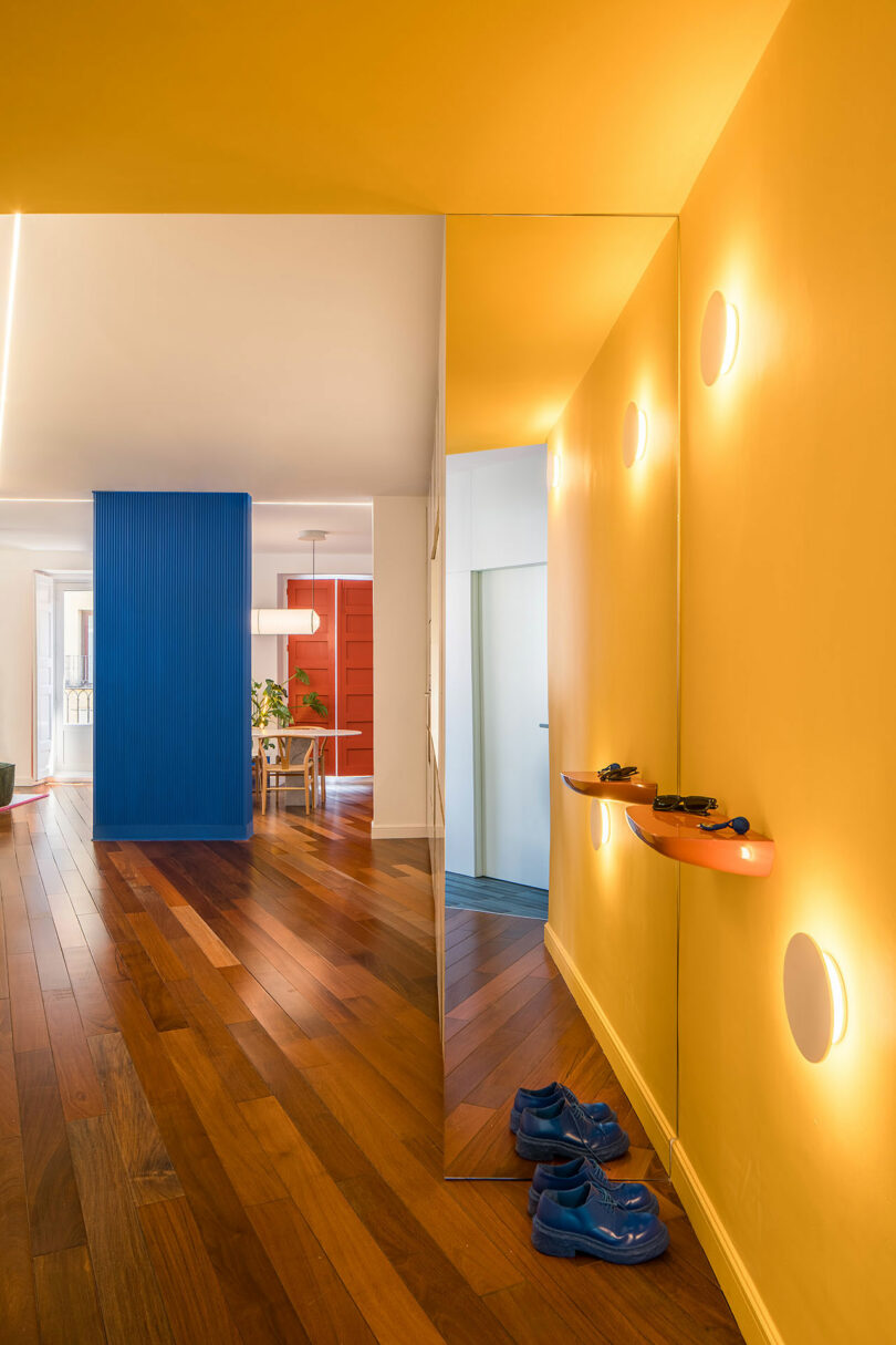 A hallway with yellow walls, a large mirror, wall-mounted shelves, blue shoes on wooden floor, and a view of a modern living space with blue and red accent walls.