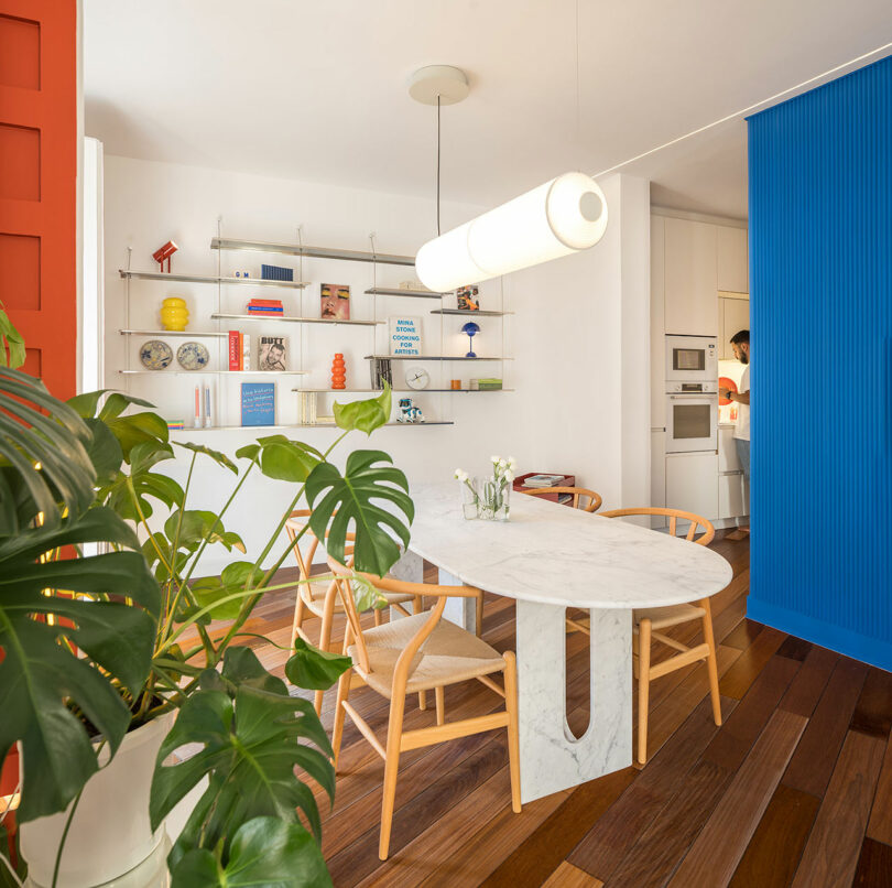 Modern dining area with a marble table, wooden chairs, houseplant, open shelving with decor, and a person in the adjacent kitchen, separated by a blue partition.